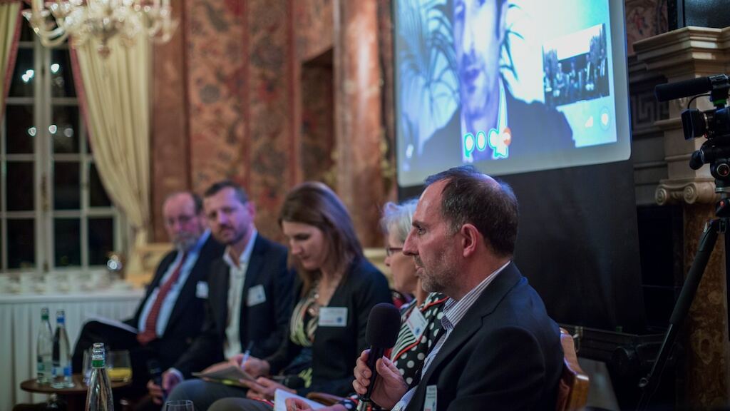 A panel of speakers seated in front of a screen showing a participant joining by video