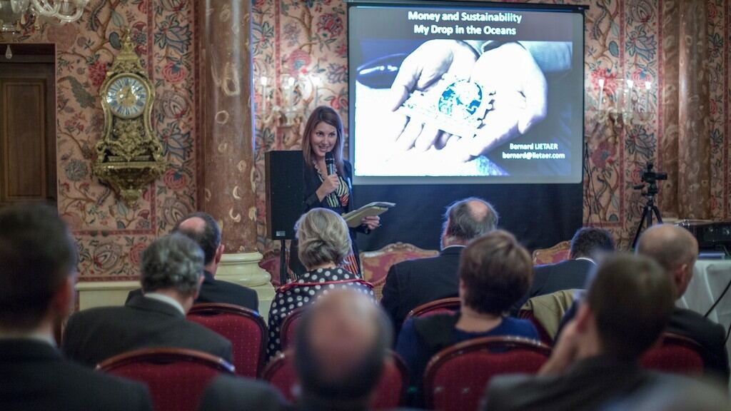 A speaker presents a slide titled Money and Sustainability, My Drop in the Oceans, to a seated audience in a panelled salon